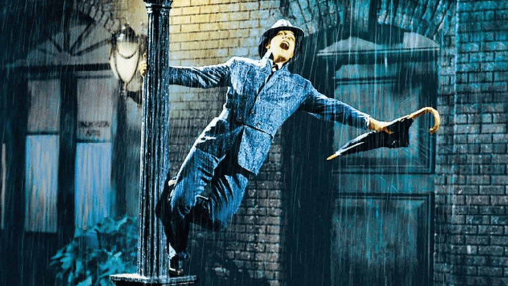Um homem de terno e chapéu segura um guarda-chuva enquanto se pendura em um poste de luz, cantando e sorrindo debaixo de uma forte chuva, em uma rua de paralelepípedos.