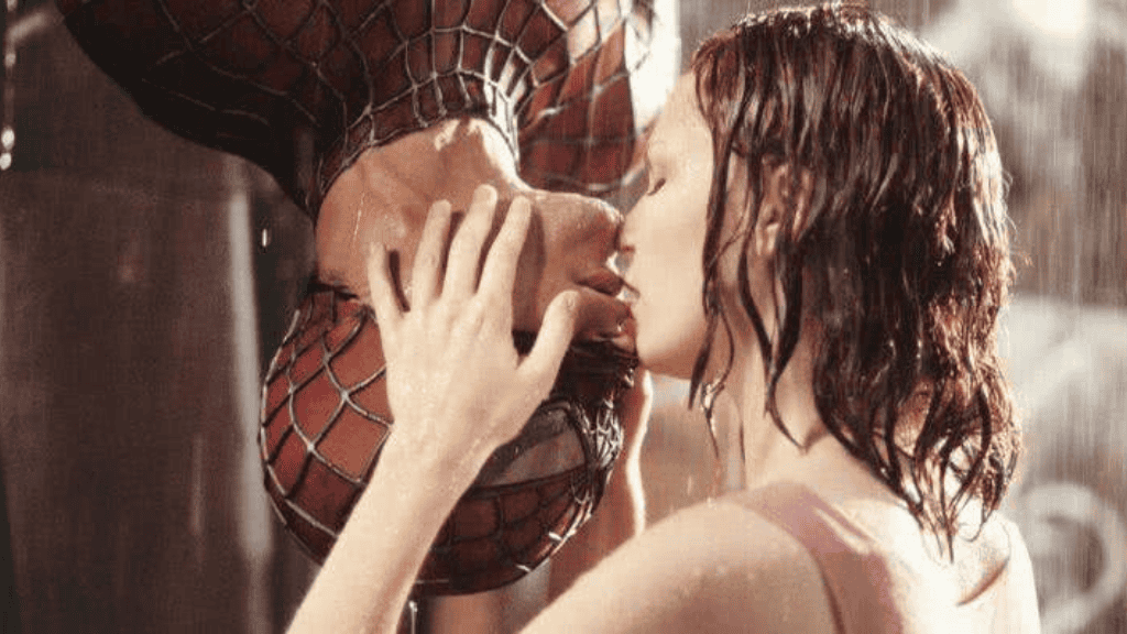 Mulher de cabelos molhados beija um homem mascarado pendurado de cabeça para baixo, enquanto a chuva cai ao redor.