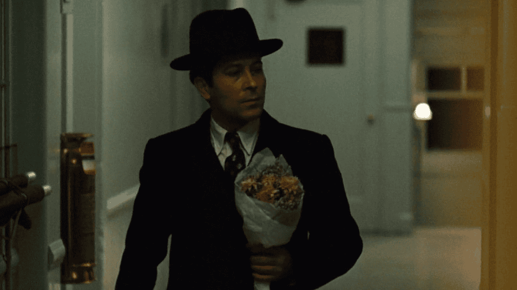 Homem de terno preto e chapéu escuro caminha por um corredor mal iluminado, segurando um pequeno buquê de flores embrulhado em papel branco. Seu rosto tem uma expressão séria e concentrada.