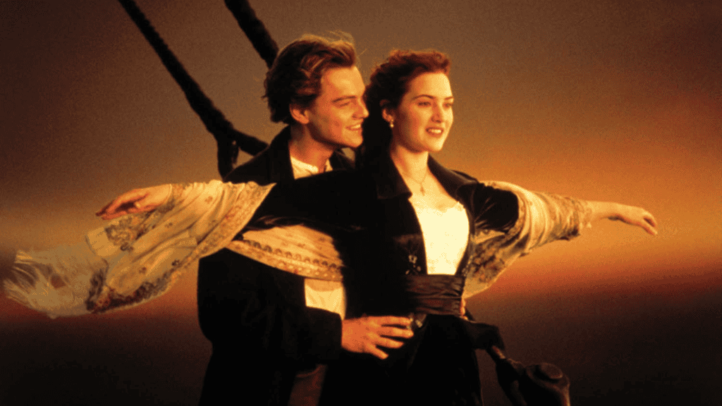 Casal jovem na proa de um navio ao pôr do sol. A mulher abre os braços enquanto o homem a segura pela cintura, sorrindo. O vento agita seu cabelo e um lenço delicado.