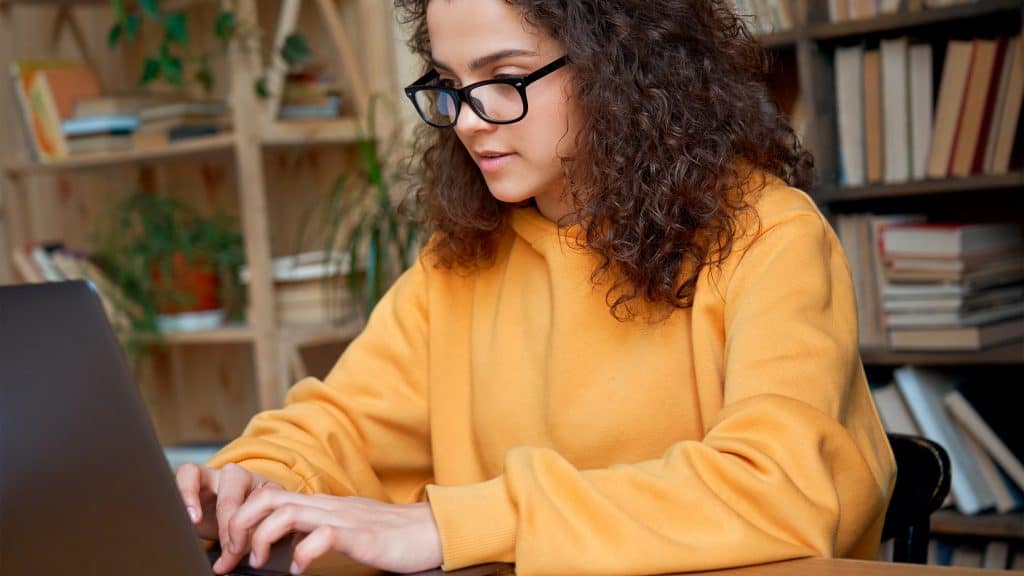 Moça usando óculos e moletom amarelo digitando em notebook enquanto estuda em casa